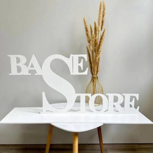 May include: White wooden sign spelling out "BASE STORE" on a white table. The letters are large and stand upright. A gold wire vase with dried wheat stalks is in the background.