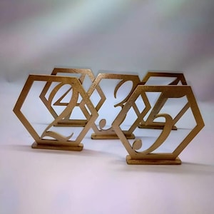 May include: Gold-colored table number holders in a hexagonal shape. Each holder features a different number, including 2, 3, 4, and 5, in an elegant script font. The holders are freestanding and suitable for events.