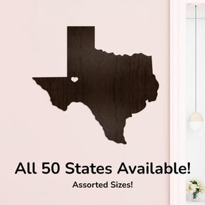 Op de afbeelding: Een donkerbruine houten uitsparing van de staat Texas, met een hartvormige uitsparing. De tekst "All 50 States Available!" en "Assorted Sizes!" wordt hieronder weergegeven. De uitsparing is op een lichtroze muur gemonteerd.