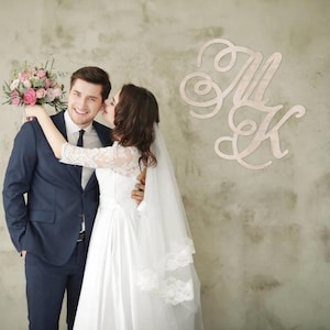 Può includere: Una coppia di sposi si abbraccia, la sposa indossa un abito da sposa bianco e un velo, lo sposo un abito blu navy. La sposa tiene in mano un bouquet di fiori rosa e bianchi. Le iniziali in legno "M K" sono montate sul muro.