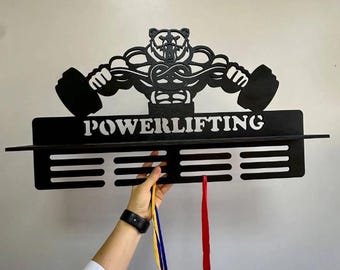 May include: Black wooden powerlifting medal display with a bear design holding a barbell. The word "POWERLIFTING" is displayed in white. The display has a shelf and slots for medals. A hand is holding medals with blue, yellow, and red ribbons.
