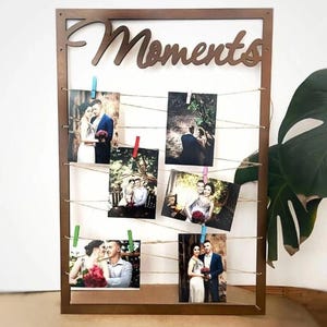 Peut inclure: Un présentoir photo en bois marron avec le mot "Moments" en écriture cursive. Plusieurs photos sont accrochées à de la ficelle avec des pinces à linge colorées. Les photos représentent des couples, probablement des photos de mariage ou de fiançailles.
