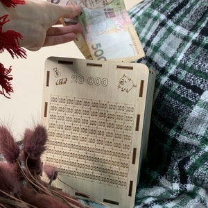 May include: A wooden piggy bank with a slot for money. The bank has a grid of squares with numbers printed on them, indicating the amount of money that can be saved. The bank also has a small pig illustration and the text "20 000" printed on it.