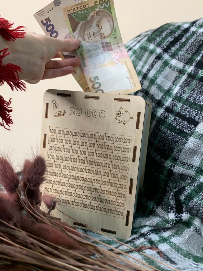 May include: A wooden piggy bank with a slot for money. The bank has a grid of squares with numbers printed on them, indicating the amount of money that can be saved. The bank also has a small pig illustration and the text "20 000" printed on it.