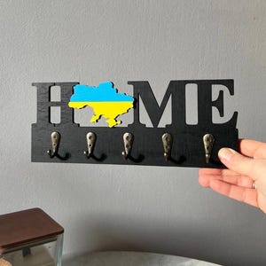 以下が含まれることがあります： 「HOME」の文字が切り抜かれた黒い木製のキーホルダー。 「O」は青と黄色のウクライナの地図に置き換えられています。 5つのブロンズ色のフックが文字の下にあります。