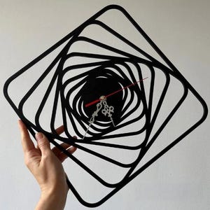 Op de afbeelding: Zwarte, geometrische wandklok met een spiraalvormig ontwerp. De klok heeft een zwarte wijzerplaat, zilveren wijzers en een rode secondewijzer. De klok wordt door een hand tegen een witte achtergrond gehouden. Het ontwerp is modern en abstract.