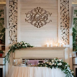 Op de afbeelding: Een bruiloft receptietafel met een wit tafelkleed en bloemstukken. Een houten bord met de initialen "M&B" is aan de muur bevestigd. De tafel is gedekt met glazen en borden, en kaarsen branden op de schoorsteenmantel.