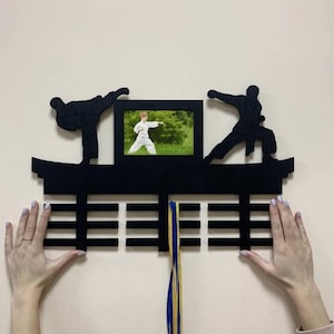 Könnte beinhalten: Schwarze Wanddekoration aus Holz mit Silhouetten von Kampfsportlern und einem gerahmten Foto einer Person in Karate-Uniform. Die Dekoration umfasst eine Reihe horizontaler Balken und ein blau-gelbes Band.