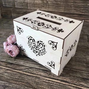 May include: A light-colored wooden card box with a slot on top and a heart-shaped cutout on the front. The box is decorated with black floral and heart-shaped designs. Pink flowers are placed next to the box.