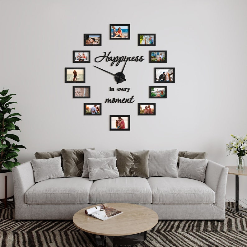 Op de afbeelding: Een zwarte fotolijstklok met de woorden "Happiness in every moment" op een witte muur. De klok is omringd door 12 kleinere zwarte fotolijsten, die elk een foto van een stel bevatten.