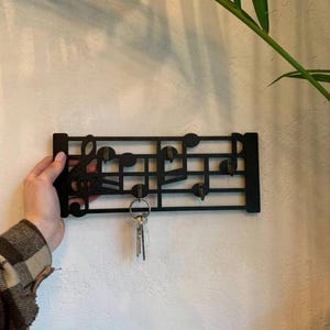 Puede incluir: Llavero de metal negro con forma de notas y símbolos musicales. El llavero presenta una clave de sol y varios ganchos para colgar llaves. Un juego de llaves cuelga de uno de los ganchos. El llavero está montado en una pared blanca.