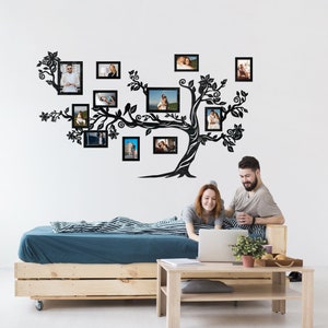 May include: A black silhouette of a tree with branches that hold multiple black picture frames. The tree is mounted on a white wall above a wooden bed frame with a blue and white striped bedspread. A couple is sitting on the bed, looking at a laptop.