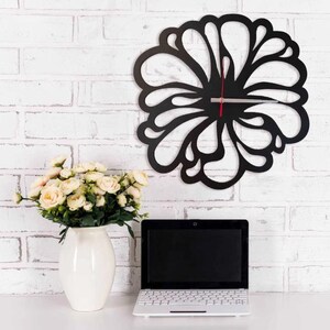 May include: A black, decorative wall clock with a floral design and a red second hand, mounted on a white brick wall. A white vase with cream roses and a laptop are on a white surface. The clock's diameter is approximately 30 cm.