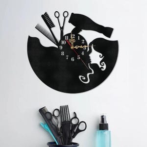 May include: Black wall clock with a salon theme. The clock features a silhouette of a woman's face, a comb, scissors, and a hairdryer. The clock hands are gold. The clock is a decorative item for a hair salon or beauty shop.