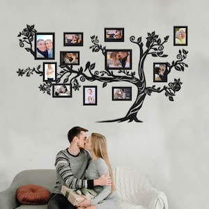 May include: Black silhouette of a tree with branches extending out, holding framed photos of various people. The tree is mounted on a white wall. A couple is sitting on a gray couch in the foreground.