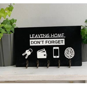 Könnte beinhalten: Ein schwarzer Holz-Schlüsselhalter mit dem weißen Text "LEAVING HOME, DON'T FORGET". Der Halter zeigt weiße Symbole von Schlüsseln, einer Brieftasche, einem Telefon und einem Gehirn, mit vier bronzenen Haken darunter.