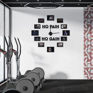 Op de afbeelding: Een wandklok met een fitnessthema. De klok heeft een zwart frame met de woorden "NO PAIN NO GAIN" en verschillende kleine ingelijste foto's. De wijzers van de klok zijn zwart en de achtergrond is een witte muur.