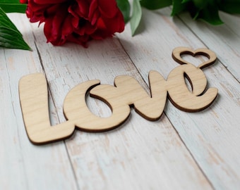 May include: Wooden "Love" sign with a heart-shaped design on the letter "e". The sign is light brown and sits on a white wooden surface, with a red flower and green leaves in the background. A decorative item for home decor.