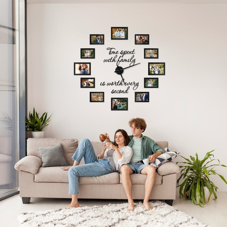 Op de afbeelding: Een zwarte fotoklok met de tekst "Time spent with family is worth every second" op een witte muur. De klok is omringd door zwarte fotolijsten met foto's van een gezin. Een stel zit op een beige bank voor de klok.