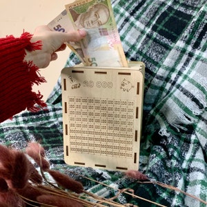 May include: A wooden piggy bank with a slot for coins. The front of the bank has a grid with numbers 100 and 200 printed on it. The bank also has the text "20 000" printed on it. A hand is putting a 50 hryvnia bill into the slot.