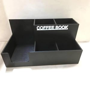 May include: A black wooden coffee organizer with multiple compartments. The organizer has the words "COFFEE BOOK" in white lettering on the front. It features a large open area on the left and several smaller sections for storage.
