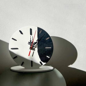 Puede incluir: Un moderno reloj de escritorio de dos tonos con una esfera blanca y negra. El reloj tiene forma redonda con marcadores de hora negros y manecillas negras, y un segundero rojo. El reloj se apoya en una base blanca.