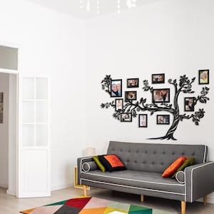 May include: A black metal tree silhouette wall decor with multiple photo frames holding family photos. The tree is mounted on a white wall above a gray sofa with pillows. The room has a white door and a colorful rug.