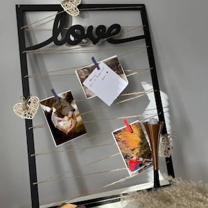 Puede incluir: Un expositor de fotos con marco negro con la palabra "love" en cursiva. Las fotos están sujetas a una cuerda con pinzas. Corazones decorativos tejidos y un candelabro de cobre añaden encanto. Las fotos muestran una pareja y una nota manuscrita.