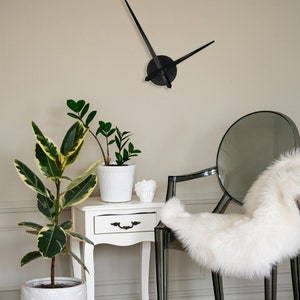 Op de afbeelding: Een witte bijzettafel met een lade en een witte potplant met groene bladeren. Op de achtergrond staat een doorzichtige plastic stoel met een witte nep bontdeken. Aan de muur hangt een zwarte klok met lange wijzers.