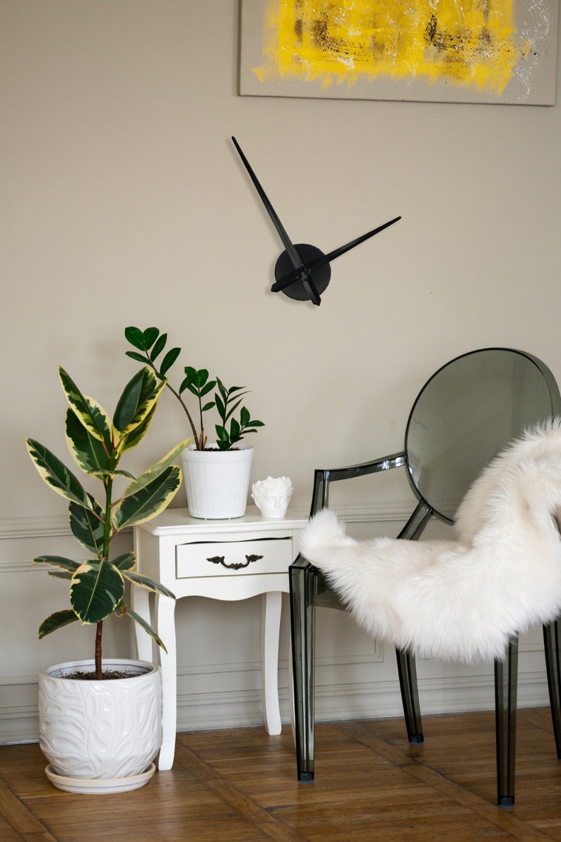 Op de afbeelding: Een witte bijzettafel met een lade en een witte potplant met groene bladeren. Op de achtergrond staat een doorzichtige plastic stoel met een witte nep bontdeken. Aan de muur hangt een zwarte klok met lange wijzers.