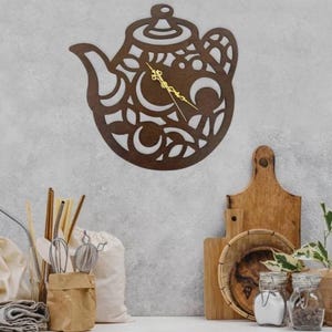 May include: A brown wooden teapot-shaped wall clock with gold-colored hands. The clock has a decorative, openwork design and is mounted on a light gray wall. Kitchen decor is visible in the background, including wooden cutting boards and jars.