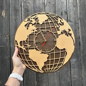 May include: A wooden world map clock with a dark brown background and a light brown globe cut-out. The clock hands are red and the numbers are Roman numerals. The clock is round and has a diameter of approximately 12 inches.