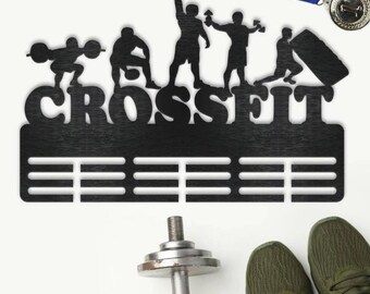 May include: Black metal CrossFit medal display with silhouettes of athletes. The word "CROSSFIT" is cut out above horizontal bars for hanging medals. A dumbbell, green athletic shoes, and a medal with a blue and yellow ribbon are also visible.