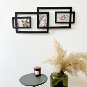 Puede incluir: Un marco de fotos negro y geométrico montado en la pared con tres aberturas para fotos. El marco contiene tres fotos de un bebé y manos. Un jarrón verde con flores secas y una pequeña mesa con una vela están en primer plano.