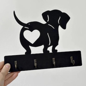 May include: Black metal dachshund key holder featuring a heart-shaped cutout. The dog silhouette sits above a rectangular base with four key hooks. Designed for wall mounting, it's a charming home accessory.