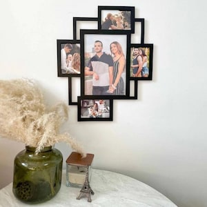 Könnte beinhalten: Ein schwarzer Mehrfach-Fotorahmen mit verschiedenen Familienfotos an einer weißen Wand. Der Rahmen hat ein einzigartiges geometrisches Design. Eine grüne Vase mit getrockneten Pflanzen, eine Kerze und eine Eiffelturm-Figur stehen auf einem Tisch im Vordergrund.