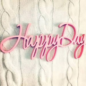 May include: A pink wooden sign with the words "Happy Day" in a cursive font. The sign is set against a textured, off-white cable knit background. The letters are a gradient of pink hues.