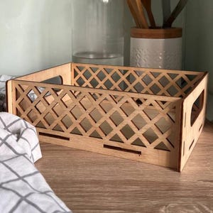 Puede incluir: Una caja de almacenamiento rectangular de madera con un diseño de celosía en los lados. La caja tiene asas recortadas y está hecha de madera de color claro. El patrón de celosía está hecho de rombos.