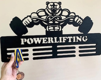 May include: Black wooden powerlifting medal holder featuring a bear lifting weights. The word "POWERLIFTING" is displayed in white. The holder has three horizontal bars for hanging medals. A hand holds a medal and a ribbon.