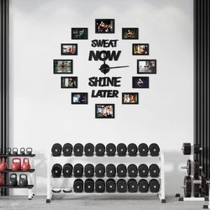 Op de afbeelding: Een home gym-opstelling met een wandklok met de tekst "SWEAT NOW SHINE LATER" en ingelijste foto's. Daaronder een rek met dumbbells en kettlebells. Fitness-thema decor.