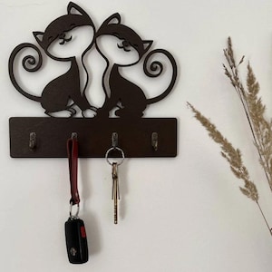 Può includere: Un portachiavi in legno marrone scuro con due sagome stilizzate di gatti. Il portachiavi ha una base rettangolare con quattro ganci. Chiavi e una chiave dell'auto sono appese ai ganci. Il portachiavi è montato su una parete bianca.