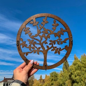 May include: A wooden key holder featuring a tree design within a circular frame. The brown-colored holder has a tree silhouette with leaves and hooks for keys. The background is a bright blue sky.