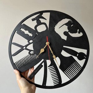 May include: Black, circular wall clock with barber-themed cutouts. The clock features scissors, combs, a razor, and a barber's pole design. The clock hands are gold with a red second hand. The clock is held by a hand.