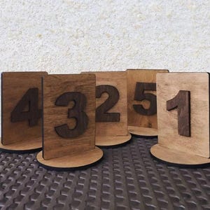 May include: Wooden table numbers 1 through 5. Each number is a dark brown, raised numeral on a light brown rectangular wooden plaque. The plaques are set on round, light brown bases.