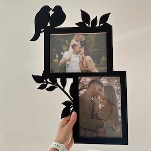 Op de afbeelding: Zwart metalen fotolijst met twee openingen voor foto's. De lijst heeft een decoratief ontwerp van vogels en bladeren. De lijst wordt door een persoon vastgehouden. De foto's binnenin tonen een gezin.