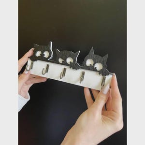 May include: A wooden key holder featuring three black cat silhouettes with large eyes, mounted on a white base. The key holder has four metal hooks for hanging keys. The design is held by a person.
