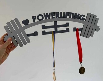 May include: A gray wooden powerlifting medal holder shaped like a barbell, with the word "POWERLIFTING" across the top. The holder has hooks for medals, with two medals hanging from it. One medal has a red ribbon, and the other has a blue and yellow ribbon.