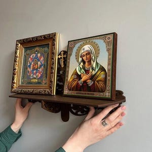 Op de afbeelding: Twee religieuze iconen op een donkerbruin houten plankje. De iconen hebben gedetailleerde kunstwerken in sierlijke lijsten. Tussen de iconen is een klein houten kruisje gemonteerd. Het plankje wordt door handen vastgehouden.