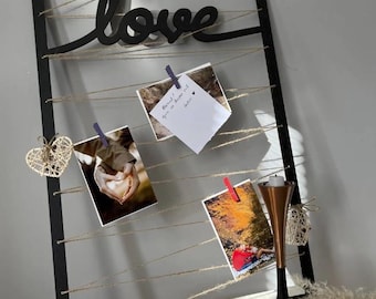 Op de afbeelding: Een zwarte houten fotodisplay met het woord "love" in cursief. De display heeft touwtjes om foto's op te hangen met wasknijpers. Decoratieve geweven harten en een koperen kandelaar dragen bij aan de decoratie.