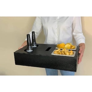 May include: A black wooden serving tray with two bottle holders and two rectangular compartments holding crisps and chips. Two dark bottles are in the bottle holders. The tray is being held by a person wearing a white shirt and blue jeans.